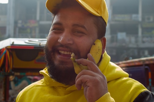 A man dressed in a yellow hoodie and cap is smiling and holding a banana to his ear, mimicking a phone. He is standing in an outdoor setting with colorful decorations and blurred buildings in the background.