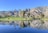 A serene landscape featuring a golf course with bright green grass, tall palm trees, and a small stone bridge over a reflective pond. In the background, rugged mountains rise under a clear, blue sky.