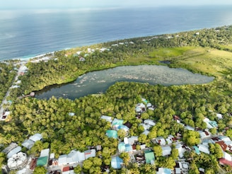 An aerial view of a coastal area featuring a large, lush green forest surrounding a serene pond. Residential houses with colorful rooftops are visible along with a shoreline bordered by the ocean.