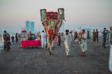 A vibrant scene on a beach with a decorated camel and people dressed in traditional attire. Several food vendors, including a stall with a bright red cloth, are present. The atmosphere is lively with groups of people interacting and enjoying the beach. Skyscrapers are visible in the hazy background, juxtaposing modernity with cultural traditions.