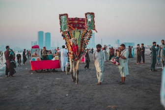 A vibrant scene on a beach with a decorated camel and people dressed in traditional attire. Several food vendors, including a stall with a bright red cloth, are present. The atmosphere is lively with groups of people interacting and enjoying the beach. Skyscrapers are visible in the hazy background, juxtaposing modernity with cultural traditions.