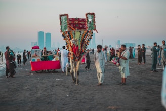 A vibrant scene on a beach with a decorated camel and people dressed in traditional attire. Several food vendors, including a stall with a bright red cloth, are present. The atmosphere is lively with groups of people interacting and enjoying the beach. Skyscrapers are visible in the hazy background, juxtaposing modernity with cultural traditions.