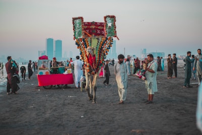 A vibrant scene on a beach with a decorated camel and people dressed in traditional attire. Several food vendors, including a stall with a bright red cloth, are present. The atmosphere is lively with groups of people interacting and enjoying the beach. Skyscrapers are visible in the hazy background, juxtaposing modernity with cultural traditions.