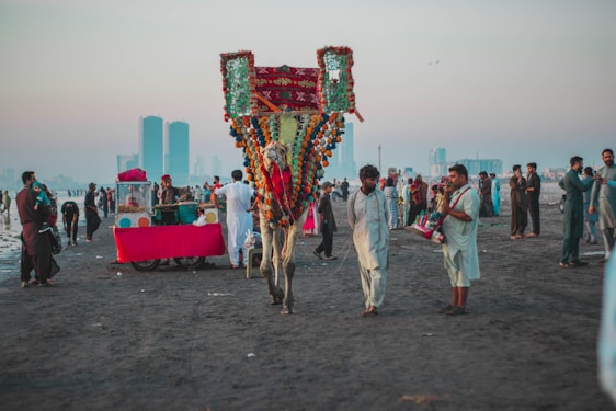 A vibrant scene on a beach with a decorated camel and people dressed in traditional attire. Several food vendors, including a stall with a bright red cloth, are present. The atmosphere is lively with groups of people interacting and enjoying the beach. Skyscrapers are visible in the hazy background, juxtaposing modernity with cultural traditions.