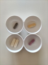 Four white bowls each containing a single capsule, arranged in a two by two grid. The capsules are of different colors: dark green, beige, white, and purple.