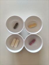 Four white bowls each containing a single capsule, arranged in a two by two grid. The capsules are of different colors: dark green, beige, white, and purple.