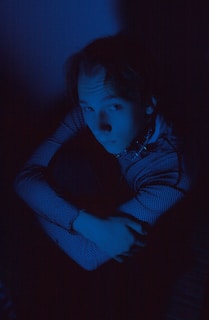 A person sits curled up, arms wrapped around their knees, in dim blue lighting. The checkered pattern on the person's shirt is visible, and their expression is contemplative.