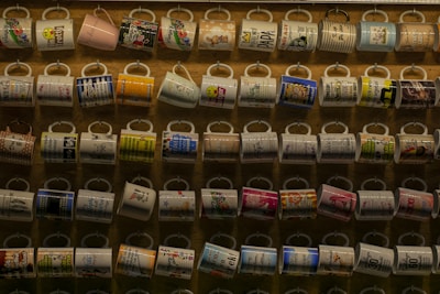 A collection of colorful mugs displayed in neat rows on a wall. Each mug features various patterns, text, and designs, showcasing a range of colors and themes. The background is a wooden surface that adds warmth to the display.