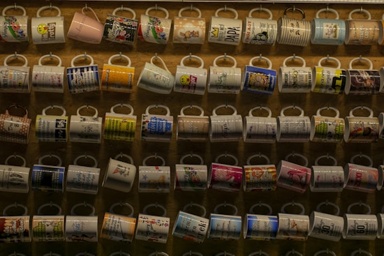 A collection of colorful mugs displayed in neat rows on a wall. Each mug features various patterns, text, and designs, showcasing a range of colors and themes. The background is a wooden surface that adds warmth to the display.