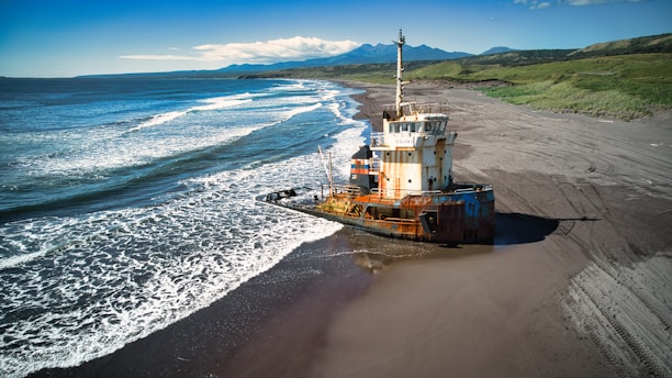 A rusted ship is stranded on a deserted sandy beach with waves gently lapping against its hull. The vast ocean stretches out to the horizon under a clear blue sky, while green hills are visible in the background, suggesting an isolated coastal environment.