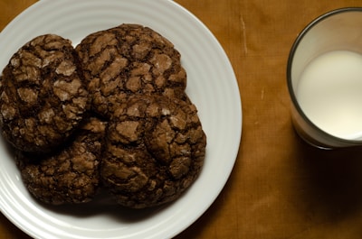 A white plate holds several chocolate cookies with a cracked surface pattern, sitting next to a glass of milk on a brown fabric surface.
