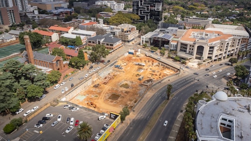 An aerial view of an urban area featuring a large construction site surrounded by roads and various buildings. The site has visible earth-moving equipment and dug-out sections. To the left, there is a red brick building with a tower, indicating an older architectural style, possibly a church or historical structure. Modern office buildings and residential complexes are visible in the background, with roads filled with numerous parked and moving vehicles.