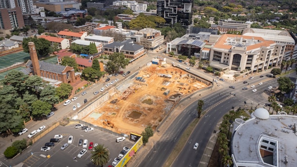 An aerial view of an urban area featuring a large construction site surrounded by roads and various buildings. The site has visible earth-moving equipment and dug-out sections. To the left, there is a red brick building with a tower, indicating an older architectural style, possibly a church or historical structure. Modern office buildings and residential complexes are visible in the background, with roads filled with numerous parked and moving vehicles.