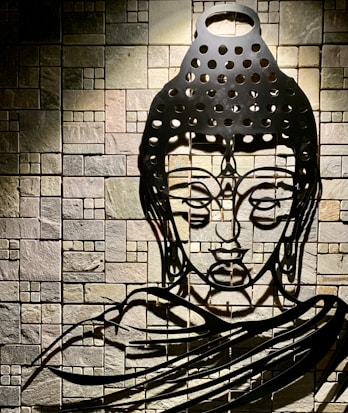 A detailed metal sculpture of a meditative face is mounted on a textured stone wall. The sculpture features various geometric cutouts casting intricate shadows, adding depth and dimension. The stone wall consists of small, square tiles in varied earthy tones.