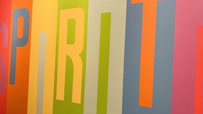 Colorful vertical stripes form the word 'SPIRIT' on a wall, with each letter in a different vibrant hue. The background features a mix of orange, yellow, green, blue, and pink shades, creating a lively and energetic composition.