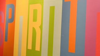 Colorful vertical stripes form the word 'SPIRIT' on a wall, with each letter in a different vibrant hue. The background features a mix of orange, yellow, green, blue, and pink shades, creating a lively and energetic composition.