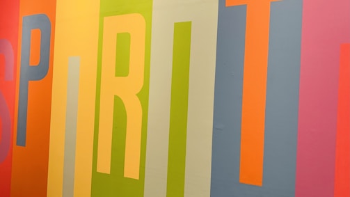Colorful vertical stripes form the word 'SPIRIT' on a wall, with each letter in a different vibrant hue. The background features a mix of orange, yellow, green, blue, and pink shades, creating a lively and energetic composition.