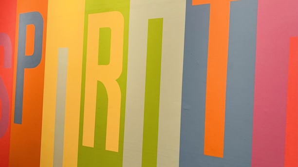 Colorful vertical stripes form the word 'SPIRIT' on a wall, with each letter in a different vibrant hue. The background features a mix of orange, yellow, green, blue, and pink shades, creating a lively and energetic composition.