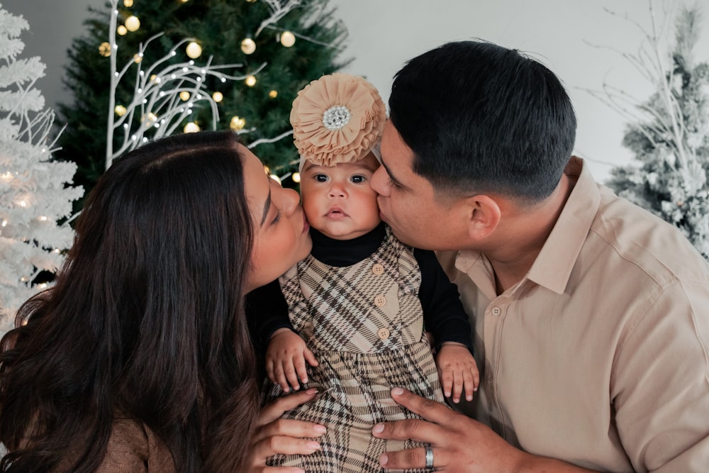 um homem e uma mulher beijando um bebê na frente de uma árvore de Natal