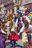 A vibrant street scene with people dressed in colorful traditional costumes, including ornate masks and pom-pom adorned headdresses. The background features a busy street lined with shops and many spectators observing the event.