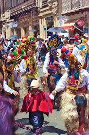 A vibrant street scene with people dressed in colorful traditional costumes, including ornate masks and pom-pom adorned headdresses. The background features a busy street lined with shops and many spectators observing the event.