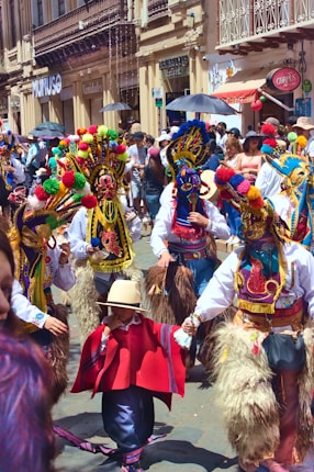 A vibrant street scene with people dressed in colorful traditional costumes, including ornate masks and pom-pom adorned headdresses. The background features a busy street lined with shops and many spectators observing the event.