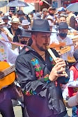 A lively street parade featuring musicians in traditional attire, with a focus on a man playing a wooden flute. The participants wear embroidered clothing and hats, and carry guitars and other instruments. The background is filled with a crowd of people, some holding umbrellas.