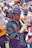 A lively street parade featuring musicians in traditional attire, with a focus on a man playing a wooden flute. The participants wear embroidered clothing and hats, and carry guitars and other instruments. The background is filled with a crowd of people, some holding umbrellas.