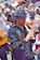A lively street parade featuring musicians in traditional attire, with a focus on a man playing a wooden flute. The participants wear embroidered clothing and hats, and carry guitars and other instruments. The background is filled with a crowd of people, some holding umbrellas.