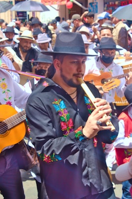 A lively street parade featuring musicians in traditional attire, with a focus on a man playing a wooden flute. The participants wear embroidered clothing and hats, and carry guitars and other instruments. The background is filled with a crowd of people, some holding umbrellas.