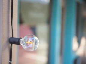 A close-up of a clear light bulb attached to a vertical wooden post with a black wire. The background is blurred but shows blue and green hues, giving a sense of an outdoor setting, possibly near a building or structure.