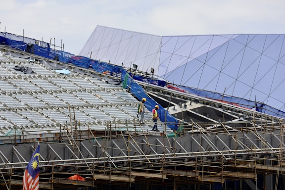 Construction workers wearing safety helmets are walking on scaffolding at a building site. The structure in progress is metallic with extensive scaffolding and temporary barriers. In the background, a modern building with angular, reflective surfaces is visible. A Malaysian flag is prominently displayed, adding a sense of national identity to the scene.