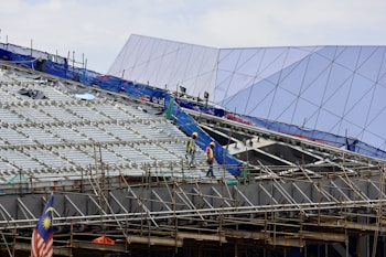 Construction workers wearing safety helmets are walking on scaffolding at a building site. The structure in progress is metallic with extensive scaffolding and temporary barriers. In the background, a modern building with angular, reflective surfaces is visible. A Malaysian flag is prominently displayed, adding a sense of national identity to the scene.