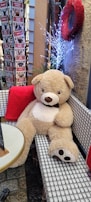 A large plush teddy bear is sitting on a checkered bench in a cozy setting. It has a light brown color with a white belly and a black bow tie. The setting includes a small round table nearby, decorated with a simple centerpiece. There is a bright red wreath hanging on the wall behind the bear and some lit decorative branches beside it, adding a festive and warm atmosphere. To the left, stacks of boxed items with French labeling are visible.