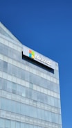 A modern glass building with a large Microsoft logo prominently displayed near the top. The sky is clear and blue, reflecting off the glass panes.