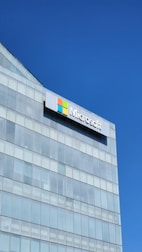 A modern glass building with a large Microsoft logo prominently displayed near the top. The sky is clear and blue, reflecting off the glass panes.