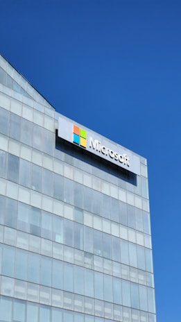 A modern glass building with a large Microsoft logo prominently displayed near the top. The sky is clear and blue, reflecting off the glass panes.
