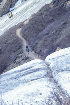 A narrow, winding path cuts through a rugged, mountainous terrain with sparse vegetation. Two hikers equipped with backpacks walk along the trail, surrounded by steep slopes and patches of snow and rock.