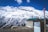 A stunning view of snow-capped mountains under a vivid blue sky, with a rustic wooden building in the foreground. A horse stands nearby surrounded by nature. There is a small graffitied structure with the words 'Save Himalayas' and a flagpole displaying a red flag.