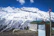A stunning view of snow-capped mountains under a vivid blue sky, with a rustic wooden building in the foreground. A horse stands nearby surrounded by nature. There is a small graffitied structure with the words 'Save Himalayas' and a flagpole displaying a red flag.