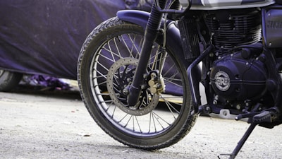 A close-up view of the front wheel and suspension system of a motorcycle, with parts of the engine visible. The background is blurred, showing a dark-colored cover on what appears to be another vehicle or object.