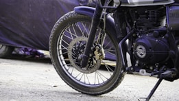 A close-up view of the front wheel and suspension system of a motorcycle, with parts of the engine visible. The background is blurred, showing a dark-colored cover on what appears to be another vehicle or object.