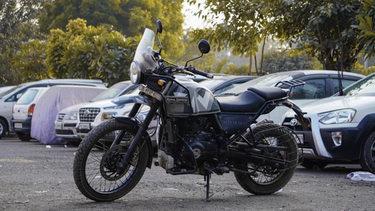 A Royal Enfield motorcycle is parked in an outdoor parking lot surrounded by several cars. The bike is predominantly black with a rugged design, featuring thick tires and a windshield. In the background, there are trees and greenery.