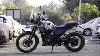 A motorcycle is parked in an outdoor setting, surrounded by several cars. The motorcycle is a Royal Enfield with a rugged design, indicating it may be used for off-road or adventure riding. The background includes trees and a partially visible building, adding to the urban environment.