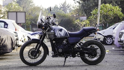 A motorcycle is parked in an outdoor setting, surrounded by several cars. The motorcycle is a Royal Enfield with a rugged design, indicating it may be used for off-road or adventure riding. The background includes trees and a partially visible building, adding to the urban environment.