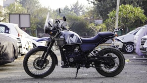 A motorcycle is parked in an outdoor setting, surrounded by several cars. The motorcycle is a Royal Enfield with a rugged design, indicating it may be used for off-road or adventure riding. The background includes trees and a partially visible building, adding to the urban environment.