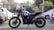 A motorcycle is parked in an outdoor setting, surrounded by several cars. The motorcycle is a Royal Enfield with a rugged design, indicating it may be used for off-road or adventure riding. The background includes trees and a partially visible building, adding to the urban environment.