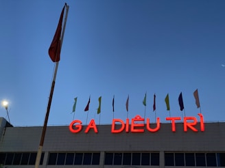 The image features a building facade with a large, red neon sign displaying the words 'GA DIÊU TRÌ' against a clear evening sky. Above the sign, there are several small flags in different colors attached to poles. A large pole with a red flag is prominently visible on the left side of the image.