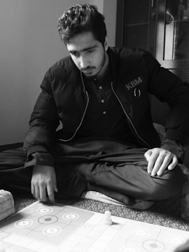 A person with a focused expression is sitting cross-legged on the floor, playing a board game. The person is wearing a dark jacket with the letters 'KBM' on it, and there is a captured moment of concentration as they engage with the game.