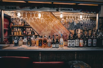 A bar counter with a wide variety of bottles lined up, including whiskey and sake. Above the counter, several glasses hang upside down from a rack, and there are bright lights illuminating the area. Shelves in the background display more bottles, and there are signs featuring brands like Coca-Cola and Kingdom Brewery.